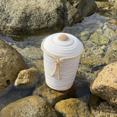 Hvit urne i plantefiber som står i strandkanten ved sjøen.