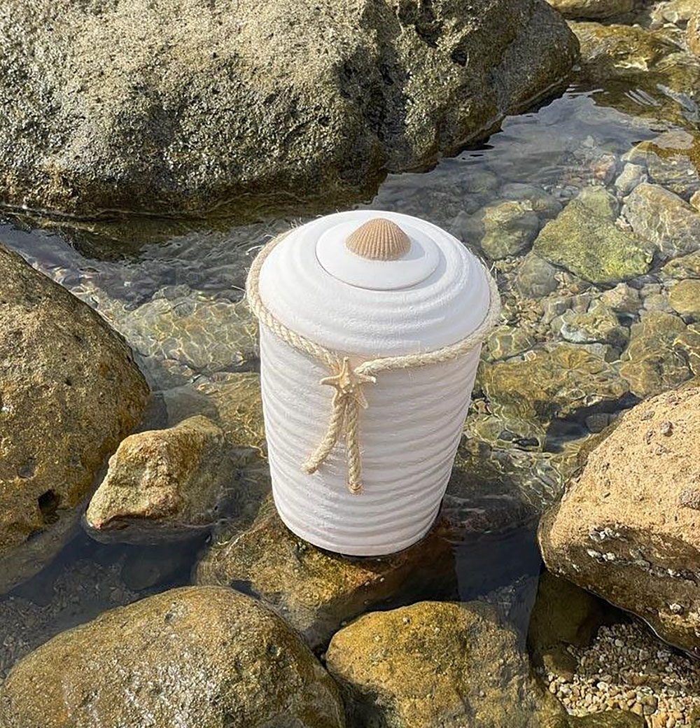 urne-plantefiber-plassert-i-vannet-blant-stein Hvit urne i plantefiber som står i strandkanten ved sjøen.