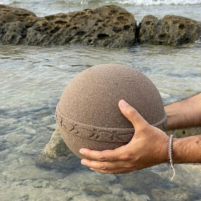 Urne i sand holdes av hender over vann fra havet.